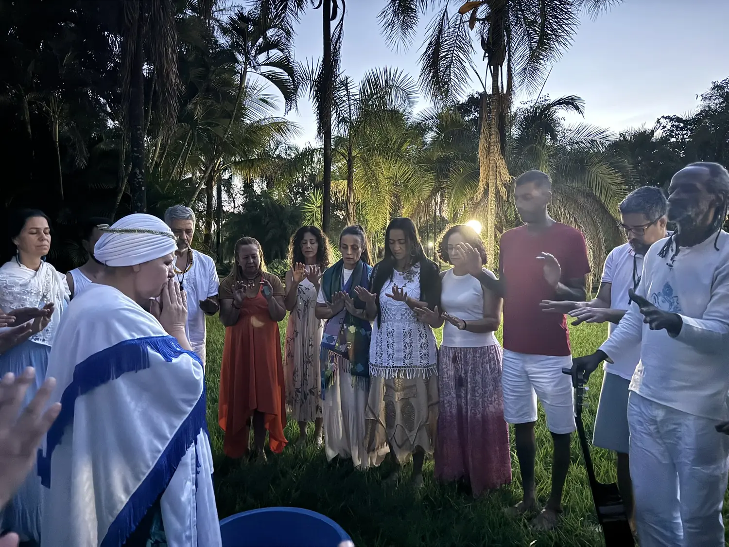 Dezenas de pessoas de branco em roda, entre palmeiras, no fim da tarde
